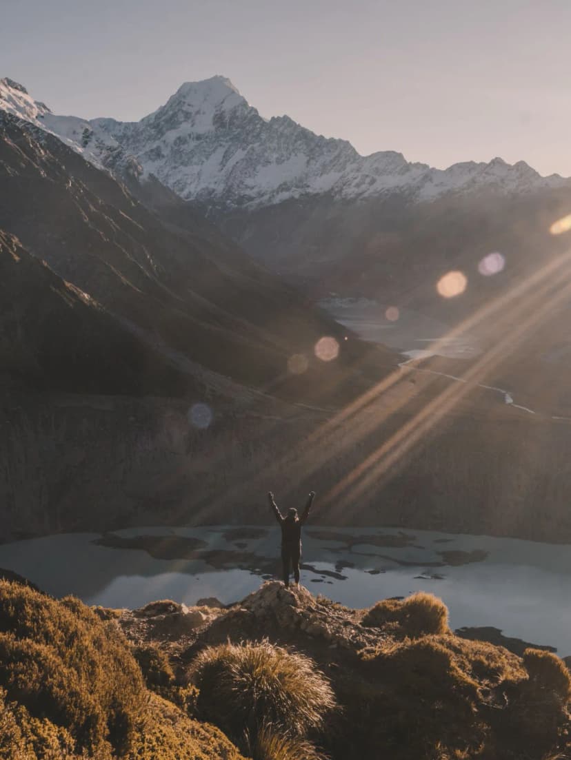 Escursionista con braccia alzate davanti al lago Mueller e alla vetta innevata di Aoraki/Mount Cook al tramonto, Nuova Zelanda.